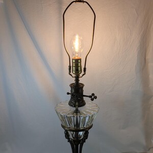 Vintage Egyptian Revival table lamp illuminated, showing ornate bronze detailing, clear glass font, and dark marble base against a light backdrop.