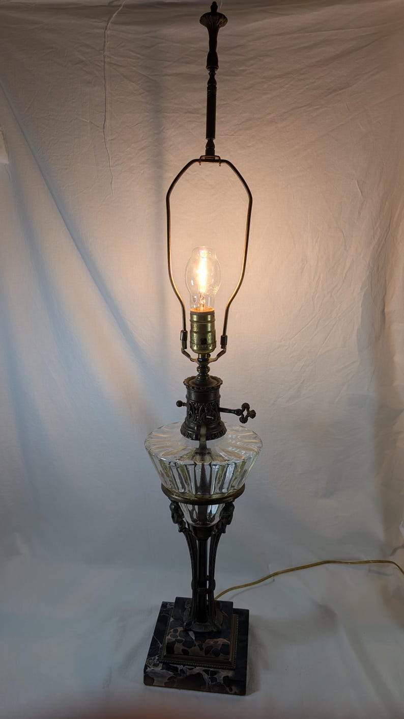 Vintage Egyptian Revival table lamp illuminated, showing ornate bronze detailing, clear glass font, and dark marble base against a light backdrop.