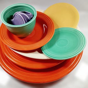 May include: A stack of four Fiestaware dinner plates in various colors, including red, yellow, and green. The top plate is a smaller, green bowl with a purple bowl inside.