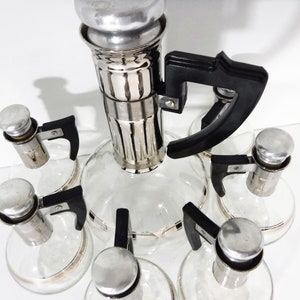 May include: A vintage glass liquor decanter set with six individual bottles. The decanter has a silver-plated top and a black plastic handle. Each bottle has a silver-plated top and a black plastic handle.