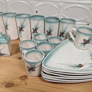 May include: A vintage ceramic dinnerware set featuring a cream-colored base with a teal rim and pine cone design. The set includes tall glasses, small cups, mugs, a sugar bowl, and triangular plates.