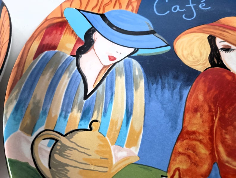 Furio Café plates displayed together: a large oval dinner plate featuring two women seated at a café table with a yellow teapot and cups, with bold, colorful artwork