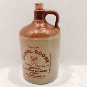 May include: A brown ceramic jug with a cork stopper and a handle. The jug is labeled "Daniel Boone Pioneer Jug Kentucky Straight Bourbon Whiskey Half Gallon."