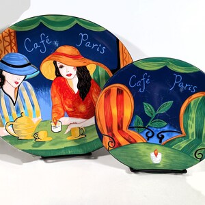 Two Sango Furio Café plates displayed together: a large oval dinner plate featuring two women seated at a café table with a yellow teapot and cups, and a smaller salad plate showing two empty striped chairs facing a candlelit table. Both plates have bold, colorful artwork with a dark blue background and “Café Paris” lettering.