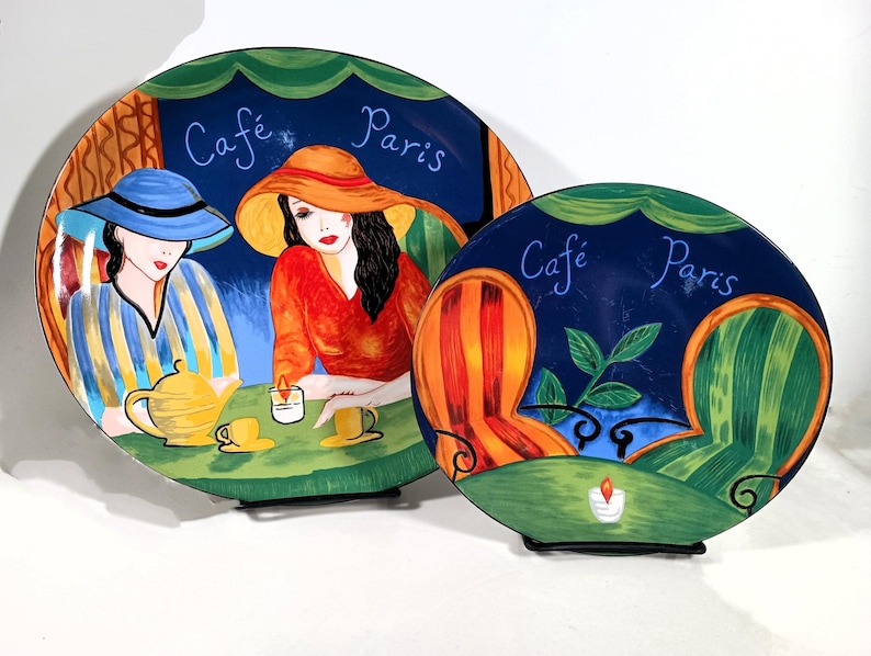 Two Sango Furio Café plates displayed together: a large oval dinner plate featuring two women seated at a café table with a yellow teapot and cups, and a smaller salad plate showing two empty striped chairs facing a candlelit table. Both plates have bold, colorful artwork with a dark blue background and “Café Paris” lettering.