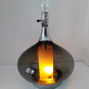 May include: A mid-century modern table lamp with a smoked glass shade and a chrome base. The lamp is lit and the light shines through the glass.