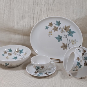 May include: A white ceramic dish set featuring a botanical design with teal, brown, and yellow leaves and flowers. The set includes a large plate, a bowl, a teacup with saucer, and a teacup with a saucer.