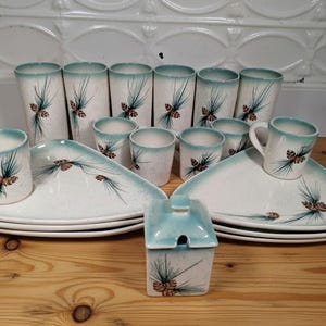 May include: A vintage ceramic dinnerware set with a white speckled finish and a teal rim. The set includes tall glasses, small cups, plates, and a sugar bowl, all decorated with a pine cone and branch design.