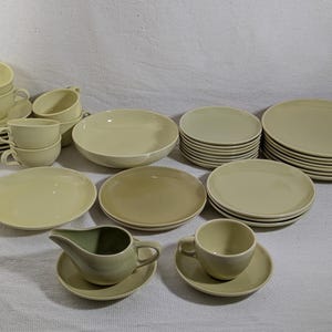May include: A collection of cream-colored ceramic dinnerware including multiple cups with saucers, various sizes of plates, a large serving bowl, and a gravy boat. The set features a smooth, solid-colored finish across all pieces, with uniform shapes.