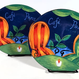 Furio Café plates displayed together: a large oval dinner plate featuring two women seated at a café table with a yellow teapot and cups, with bold, colorful artwork with
