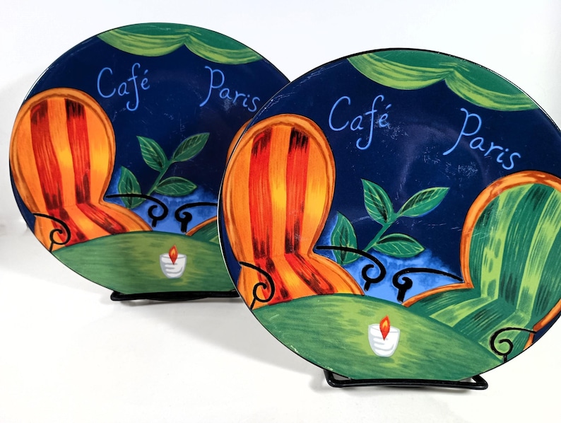 Furio Café plates displayed together: a large oval dinner plate featuring two women seated at a café table with a yellow teapot and cups, with bold, colorful artwork with