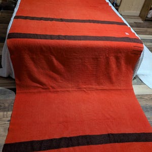 May include: A vibrant orange blanket with three dark brown stripes. The blanket is spread out on a bed, showcasing its rectangular shape and textured surface. The blanket is made of a thick material, perfect for warmth.