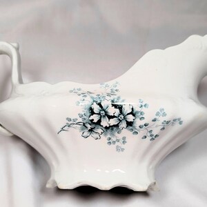 May include: White porcelain gravy boat with a curved handle and spout. The boat is decorated with a floral pattern in shades of blue and green. The base has a scalloped edge.
