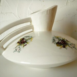 May include: White ceramic vase with a unique design featuring a wide, flat base and a tall, narrow neck. The vase has a decorative handle with three cutouts and is decorated with floral motifs in shades of green, white, and purple.