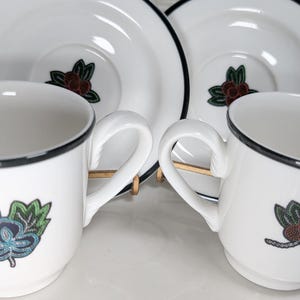 May include: White ceramic teacups and saucers with a black rim and floral design. The cups have a curved handle and a floral design on the front. The saucers also have a floral design in the center.