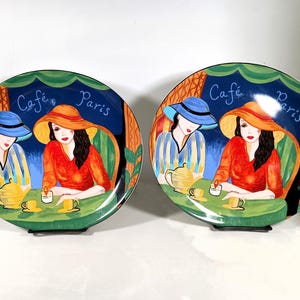 Two Sango Furio Café plates displayed together: a large oval dinner plate featuring two women seated at a café table with a yellow teapot and cups, with bold, colorful artwork with a dark blue background and “Café Paris” lettering.
