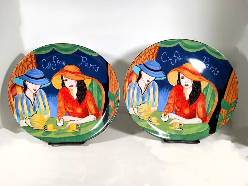 Two Sango Furio Café plates displayed together: a large oval dinner plate featuring two women seated at a café table with a yellow teapot and cups, with bold, colorful artwork with a dark blue background and “Café Paris” lettering.