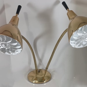 May include: A vintage brass wall-mounted lamp with two adjustable gooseneck arms. Each lamp head is gold-toned with a black accent and a white, spiral-patterned diffuser. The lamp has a round base and is designed for wall mounting.