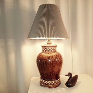 May include: A brown ceramic table lamp with a pleated gray lampshade. The lamp has a decorative pattern around the base and top. A small wooden duck figurine sits beside the lamp on a white surface.