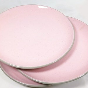 May include: Three pink ceramic dinner plates stacked on top of each other. The plates have a light gray rim.
