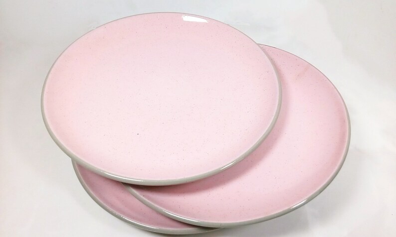 May include: Three pink ceramic dinner plates stacked on top of each other. The plates have a light gray rim.
