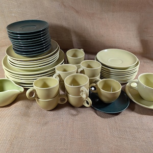 May include: A collection of vintage dishware in shades of green and cream. The set includes stacked plates, saucers, cups, and a creamer. The plates and saucers are in a light cream color, while some saucers and plates are a darker green.