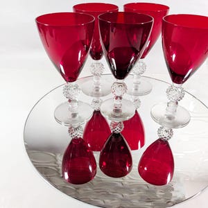 May include: Six ruby red wine glasses with crystal ball stems are arranged on a mirrored surface. The glasses have a classic design with a wide bowl and a clear base. The mirrored surface reflects the glasses, creating a symmetrical effect.