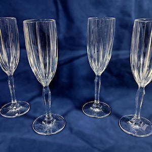 May include: Four clear glass champagne flutes with a fluted design. The flutes are arranged in a row on a blue surface.