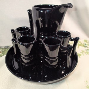 May include: A black ceramic pitcher and six matching shot glasses with handles, all on a black ceramic tray. The set is glossy and has a vintage look.