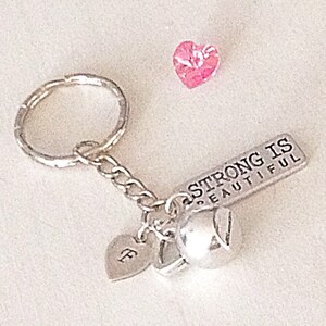May include: Silver keychain with a charm that says "Strong is Beautiful" and a small silver heart charm with the letter "F" on it. The keychain also has a small silver bell charm.