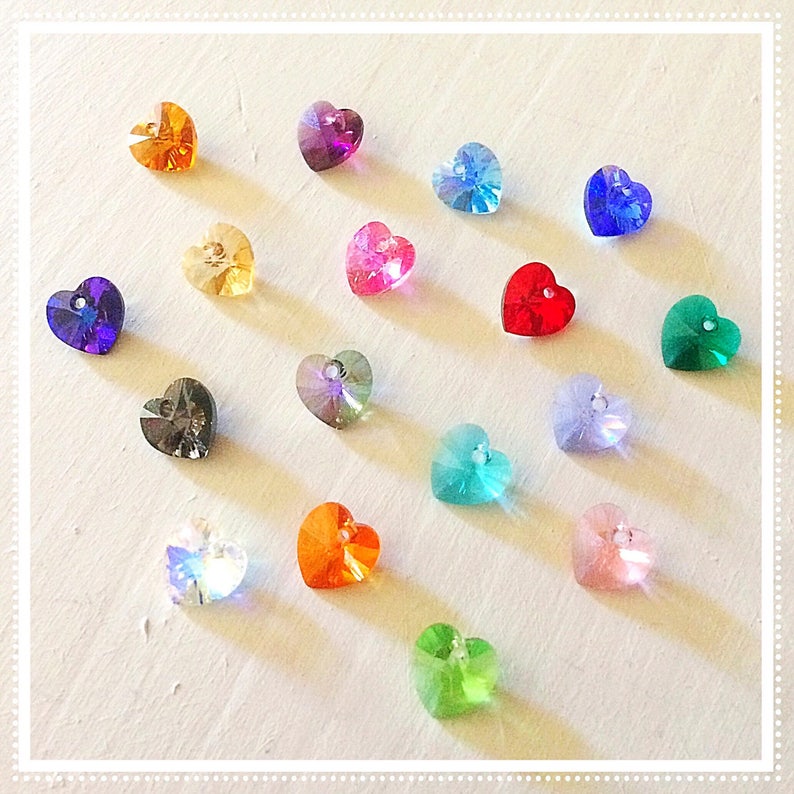 May include: Assortment of heart-shaped crystal beads in various colors, including red, blue, green, purple, orange, pink, and clear. The beads are arranged in a grid pattern on a white surface.