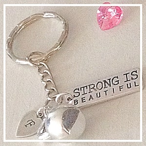 May include: Silver keychain with a charm that says "Strong is Beautiful" and two silver heart charms. One heart charm has the letter "F" engraved on it. A pink heart-shaped rhinestone is in the background.