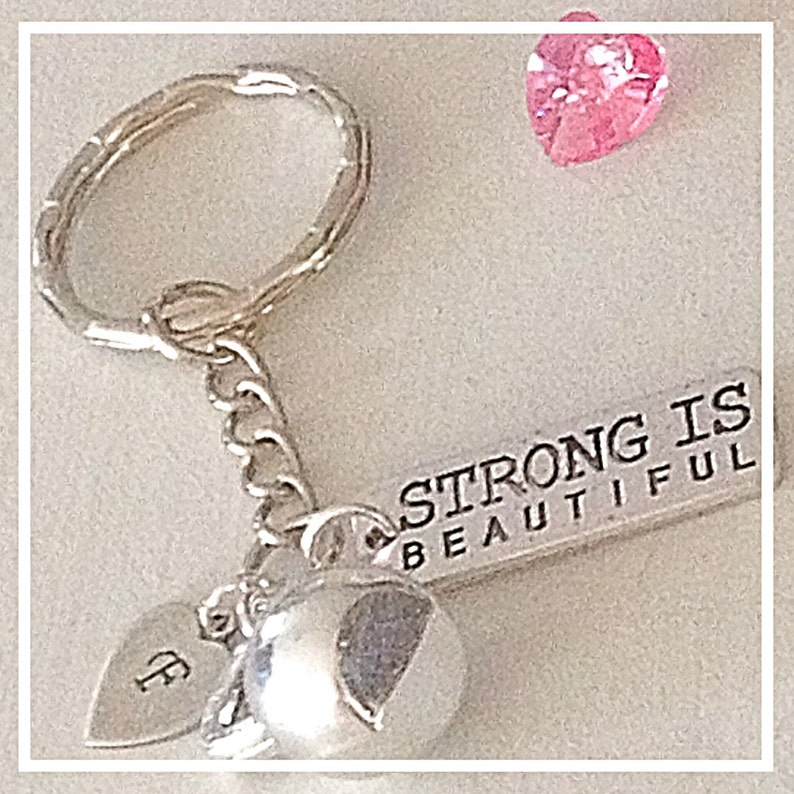 May include: Silver keychain with a charm that says "Strong is Beautiful" and two silver heart charms. One heart charm has the letter "F" engraved on it. A pink heart-shaped rhinestone is in the background.