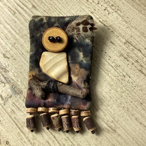 May include: A rectangular fabric brooch featuring a tie-dye pattern in shades of blue, purple, and brown. The brooch is decorated with a round wooden button, a smooth white stone, a small twig, a brown feather, and six dangling wooden beads.