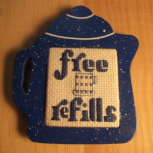 May include: A blue, teapot-shaped wooden sign with white speckles. The center features a cross-stitched design with the words "free refills" and a coffee cup illustration. The sign has a handle on the side.