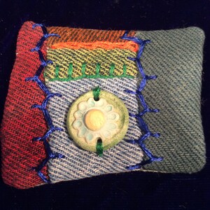 May include: A small, colorful patchwork pillow with a green and blue button in the center. The pillow is made of different fabrics, including denim, plaid, and striped fabric. The button has a flower design.