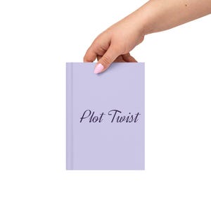 May include: A lavender-colored notebook with the words "Plot Twist" in elegant script. The notebook is held by a hand with painted nails, suggesting a focus on writing or journaling. The cover is smooth and the text is in a dark blue color.