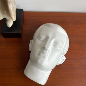 May include: A white plaster bust of a man's head, tilted slightly, with detailed facial features. The sculpture rests on a wooden surface. A second abstract sculpture is visible in the background.