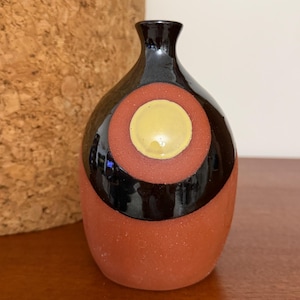 May include: A small ceramic vase with a narrow neck and a rounded body. The base is a matte terracotta color, while the upper section features a glossy black glaze with a prominent circular design containing a central pale yellow disc.