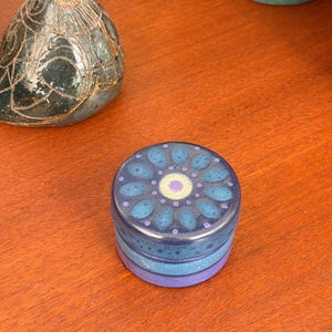 May include: A small, round ceramic lidded box featuring a dark blue base and a lid decorated with a blue and purple floral-inspired pattern. The lid displays a central light purple circle surrounded by a ring of blue petals and small dots.