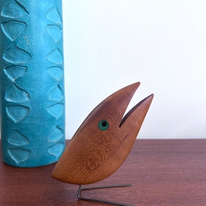 May include: A wooden bird sculpture with a green eye and an open beak. The bird is standing on a wooden surface.