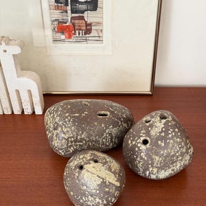 Puede incluir: Tres piedras decorativas de cerámica marrón con detalles beige moteado. Cada piedra tiene uno o más agujeros. Las piedras están dispuestas sobre una superficie de madera. Una impresión abstracta enmarcada y una escultura blanca están en el fondo.