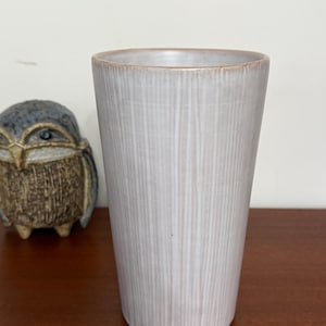 May include: A tall, white ceramic vase with a textured, wood-grain pattern. The vase has a slightly flared opening and a rounded bottom.