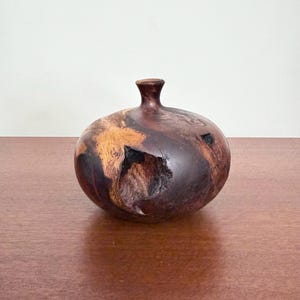 May include: A small, rounded wooden vase with a narrow neck and a wide, bulbous body. The surface features a dark, polished brown finish with natural wood grain patterns, including lighter tan sections and a deep, irregular textured hollow.