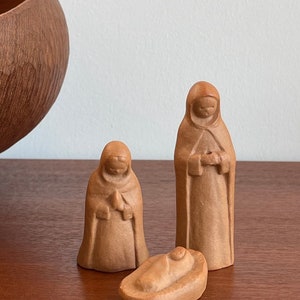 May include: A set of three light brown, smooth-textured figurines depicting a nativity scene. The collection includes two standing figures wearing hooded cloaks and one small, reclining figure wrapped in a swaddled cloth, all crafted from a matte material.