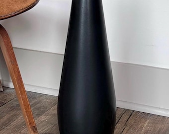 May include: A tall, black ceramic vase with a narrow opening and a rounded bottom. The vase is standing on a wooden floor.