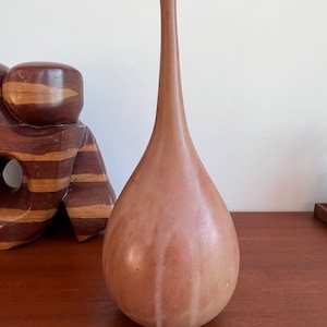 May include: A tall, slender ceramic vase with a teardrop shape. The vase is a light brown colour with a subtle glaze that creates a streaky effect.