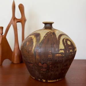 Peut inclure: Un vase en céramique marron avec un corps arrondi et un col étroit. Le vase a une surface tachetée et texturée avec des nuances de marron et de beige. Une sculpture en bois est en arrière-plan.