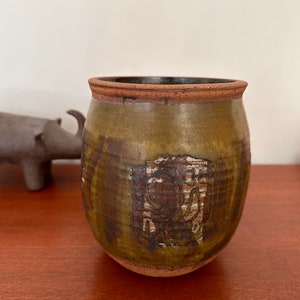 May include: A handcrafted ceramic pot with a rounded shape and a dark brown rim. The pot has a textured, olive green glaze with a rectangular, faded brown design on the front. The pot sits on a wooden surface.
