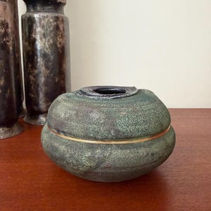 May include: A round, low-profile ceramic vessel with a textured, mottled green and dark brown finish. The piece features a horizontal gold-colored band encircling the center and a dark, circular opening at the top with a slightly uneven rim.